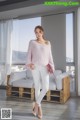 a woman standing in front of a window wearing white pants and a pink sweater