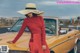 A woman in a red dress and straw hat leaning on a yellow car.