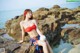 A woman in a red bikini sitting on a rock by the ocean.