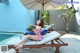 A woman in a pink dress sitting on a lounge chair by a pool.