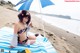 A woman in a bikini sitting on a beach under an umbrella.